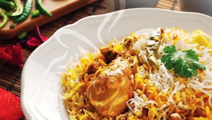 A plate of biryani with vibrant, fluffy rice and pieces of seasoned chicken garnished with fresh cilantro. A green chili pepper is visible on the plate. In the background, there are additional chilies on a wooden board.