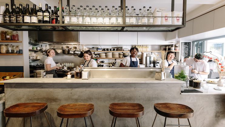 A busy, modern kitchen with five smiling chefs in white uniforms preparing food. The kitchen has a clean, industrial look with shelves of bottles and ingredients, hanging pots, and a marble counter with four wooden stools in the foreground.