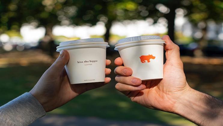 Two hands holding takeaway coffee cups against a blurred outdoor background with trees. The cup on the left reads kiss the hippo coffee, and the cup on the right features an orange hippo icon.