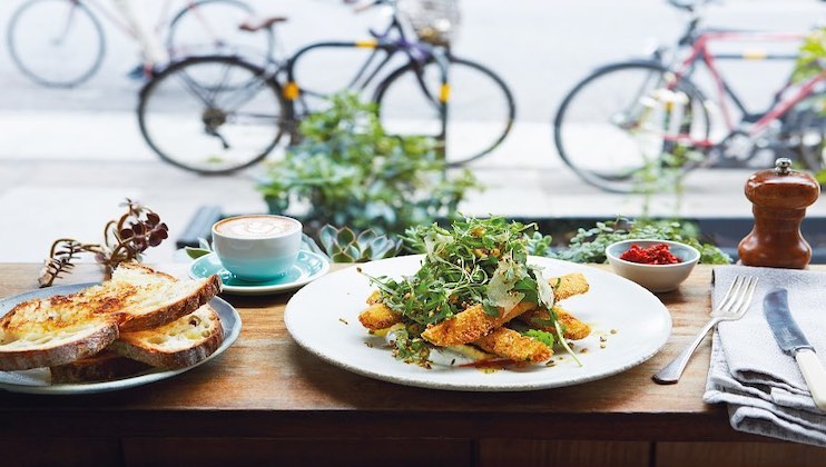 A wooden table set with breakfast items includes a plate of avocado toast garnished with arugula, a cappuccino in a turquoise cup and saucer, a side of toasted bread, red sauce in a small dish, cutlery on a napkin, and a pepper grinder. Bicycles are seen outside.