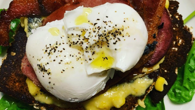 A close-up of a poached egg seasoned with cracked black pepper and sea salt, sitting atop crispy bacon and buttered toast. The toast is topped with melted cheese, and there are fresh spinach leaves at the bottom.