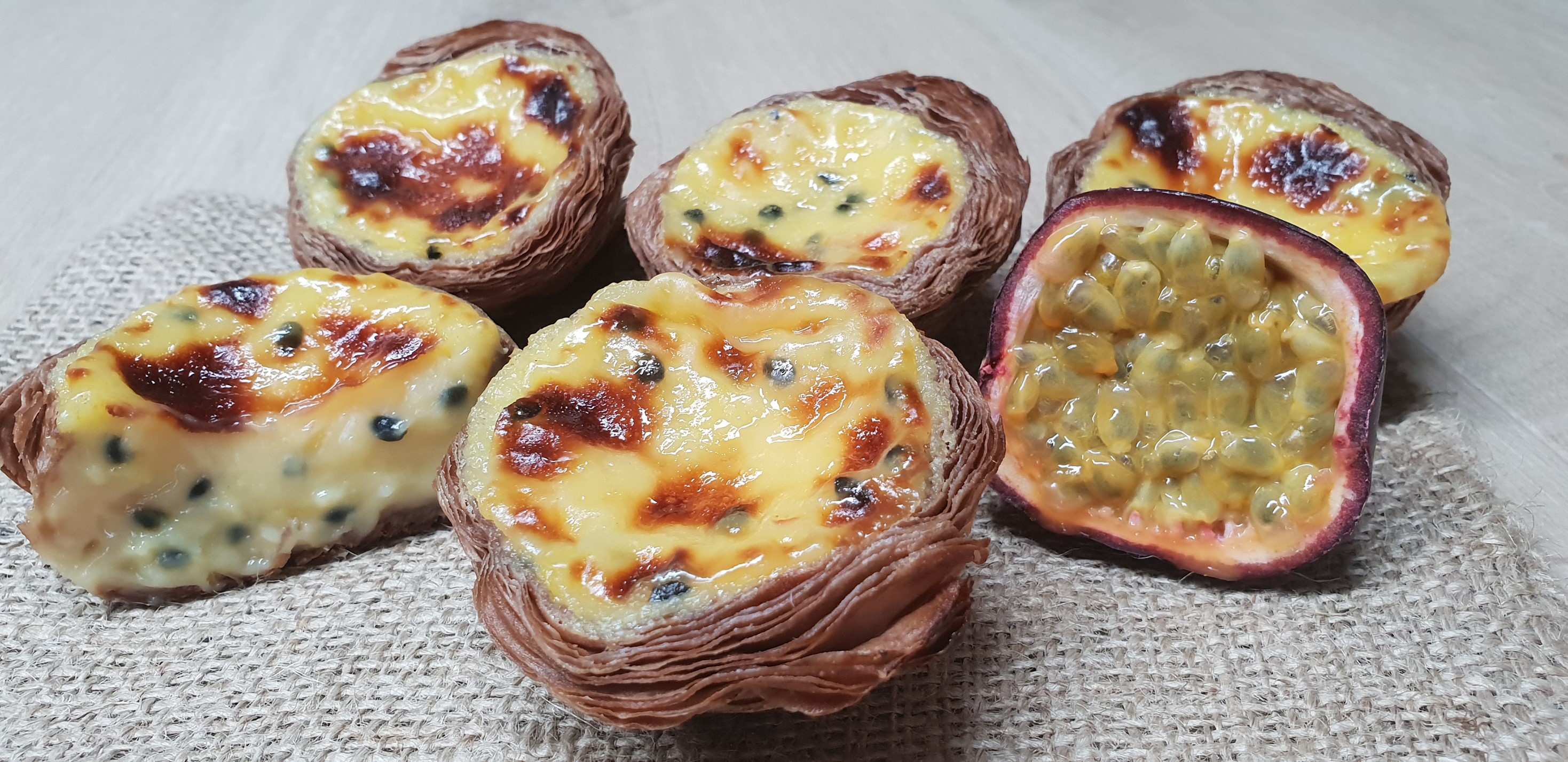 A group of freshly baked, golden-brown pastries with creamy, slightly charred tops filled with passion fruit seeds. The pastries are arranged on a burlap cloth beside a half-cut, ripe passion fruit displaying its juicy, seed-filled interior.