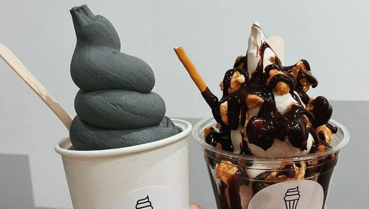 A close-up of two soft-serve ice cream desserts. The left cup holds greyish-black soft-serve with a wooden spoon. The right cup has vanilla soft-serve topped with chocolate syrup, crushed nuts, and a biscuit stick. Both are in white cups with an ice cream cone logo.