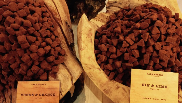 Two large wooden bowls hold piles of brown truffles. One bowl is labeled Vodka & Orange Truffles, while the other is labeled Gin & Lime Truffles. Both labels indicate the truffles contain alcohol and dairy, and they're made by Dark Sugars Chocolates.