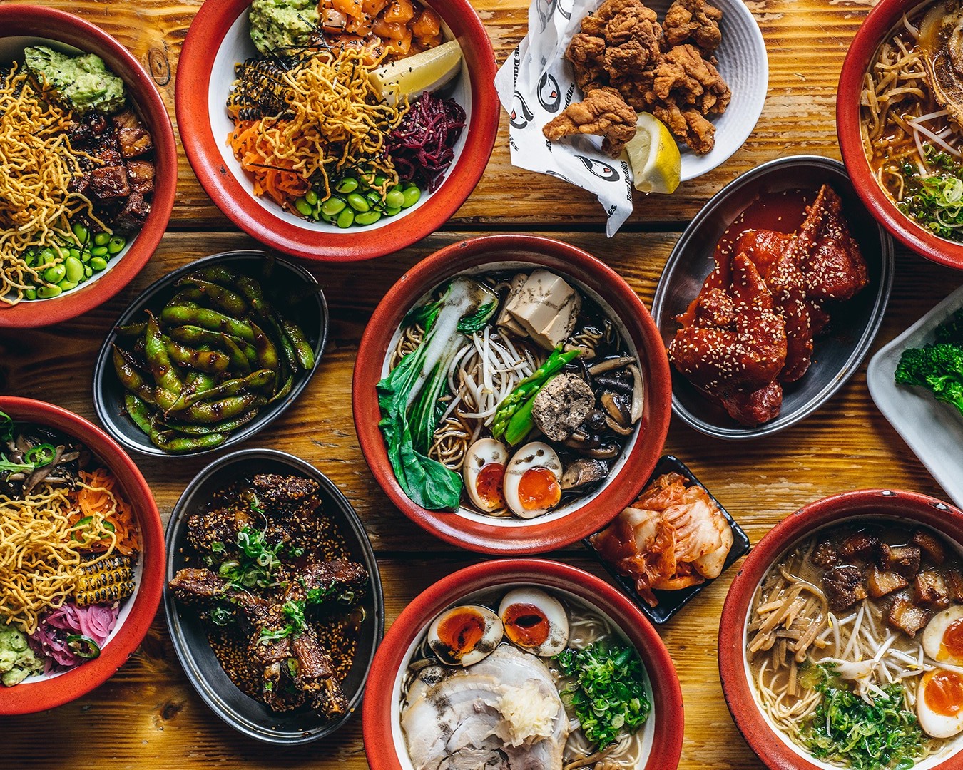 A wooden table is filled with an assortment of vibrant Asian dishes, including various types of ramen, edamame, fried chicken, seaweed salad, and other side dishes. Each bowl and plate showcases colorful ingredients, arranged in an appetizing manner.