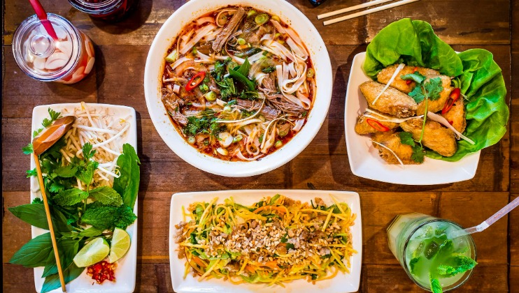 A vibrant assortment of Vietnamese dishes is displayed on a wooden table. The spread includes a bowl of noodle soup with various toppings, a small plate of fresh spring rolls, a plate of papaya salad with crushed peanuts, fresh herbs, and a drink with a green hue.