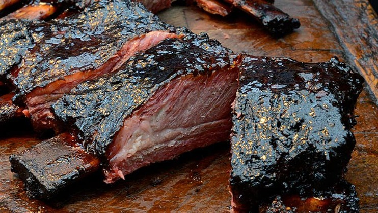 Close-up of grilled BBQ ribs sliced on a wooden cutting board. The ribs are glazed with a dark, glossy barbecue sauce and have charred edges. The meat appears tender and juicy, with a rich smoky texture.