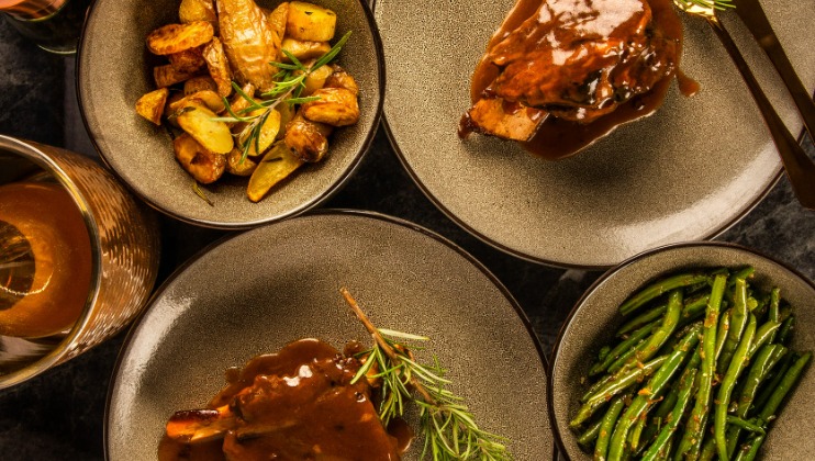 A top-down view of a meal with three plates. One plate contains roasted potatoes garnished with herbs, another has a meat dish with sauce and herbs, and the third plate has cooked green beans. A drink with a lemon slice is visible on the side.