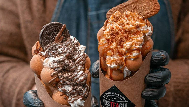 Two hands holding bubble waffles in cardboard cones. The left waffle is topped with whipped cream, crushed Oreo cookies, and a whole Oreo. The right waffle has whipped cream, caramel drizzle, crumbled cookie, and a whole cookie. A Nostalgia logo is visible on the cones.