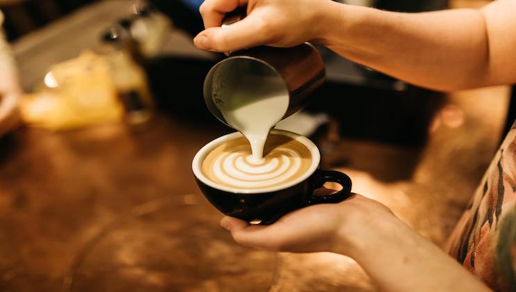 A person is pouring steamed milk from a pitcher into a cup of coffee, creating latte art with a beautiful pattern. The coffee cup is black and the background is a blurred indoor setting with warm lighting.