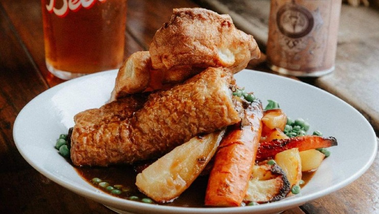 A plated meal featuring a golden-brown roasted meat with a crispy exterior, surrounded by peas, roasted potatoes, carrots, and topped with Yorkshire puddings. A glass of amber-colored drink and a can with a design are visible in the background on a wooden table.