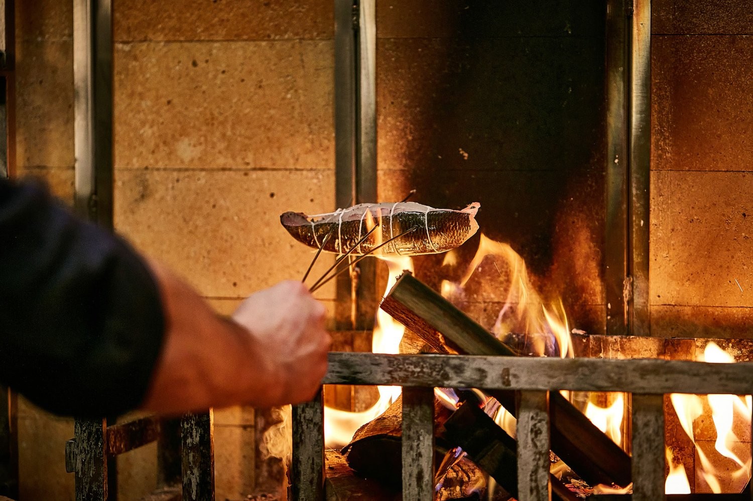 A person roasts a fish over a wood-burning fire. The fish is held by a metal skewer, and flames and glowing embers are visible beneath it. The scene takes place in front of a brick or tile backdrop.