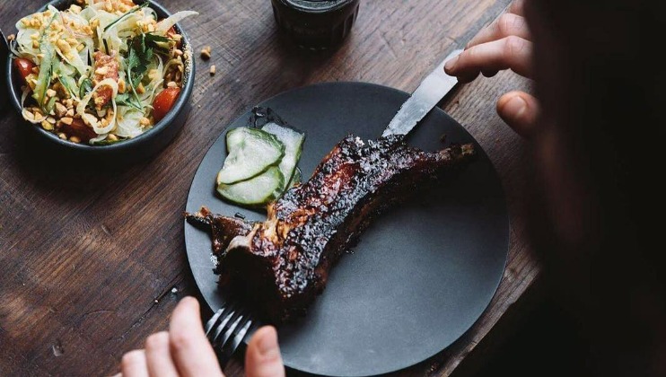 A person is cutting into a barbecued rib on a black plate with a knife and fork on a wooden table. Next to the plate is a bowl of mixed salad with tomatoes and nuts, and a drink in a glass.