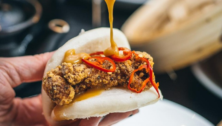 A close-up of a hand holding a steamed bun filled with fried chicken, sliced red chili peppers, and drizzled with a creamy orange sauce. The background is blurred, highlighting the focus on the appetizing dish.