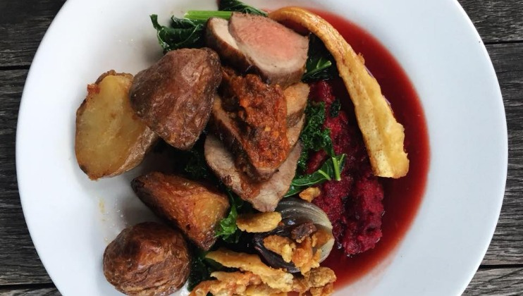 A plate of food featuring three pieces of roasted meat on top of vegetables, accompanied by roasted potatoes, a vibrant red sauce, and crispy garnish. The dish is presented on a white plate against a wooden table background.