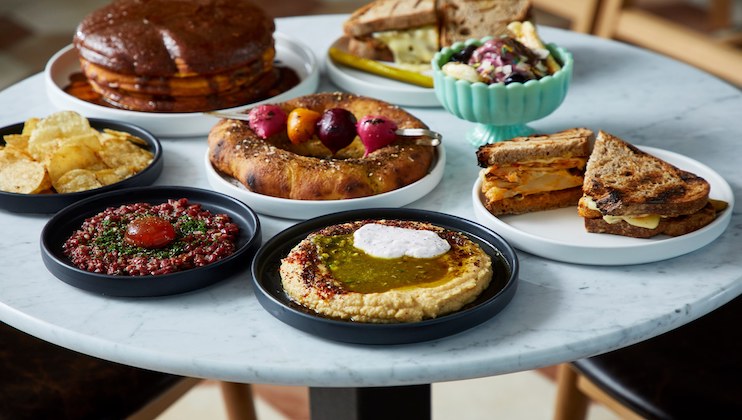 A round white table is filled with various dishes. There are dips with seasoning, a stack of pancakes, grilled sandwiches, a bowl of salad, a ring bread with skewered vegetables, and a plate of potato chips. The dishes have vibrant colors and contrasting textures.