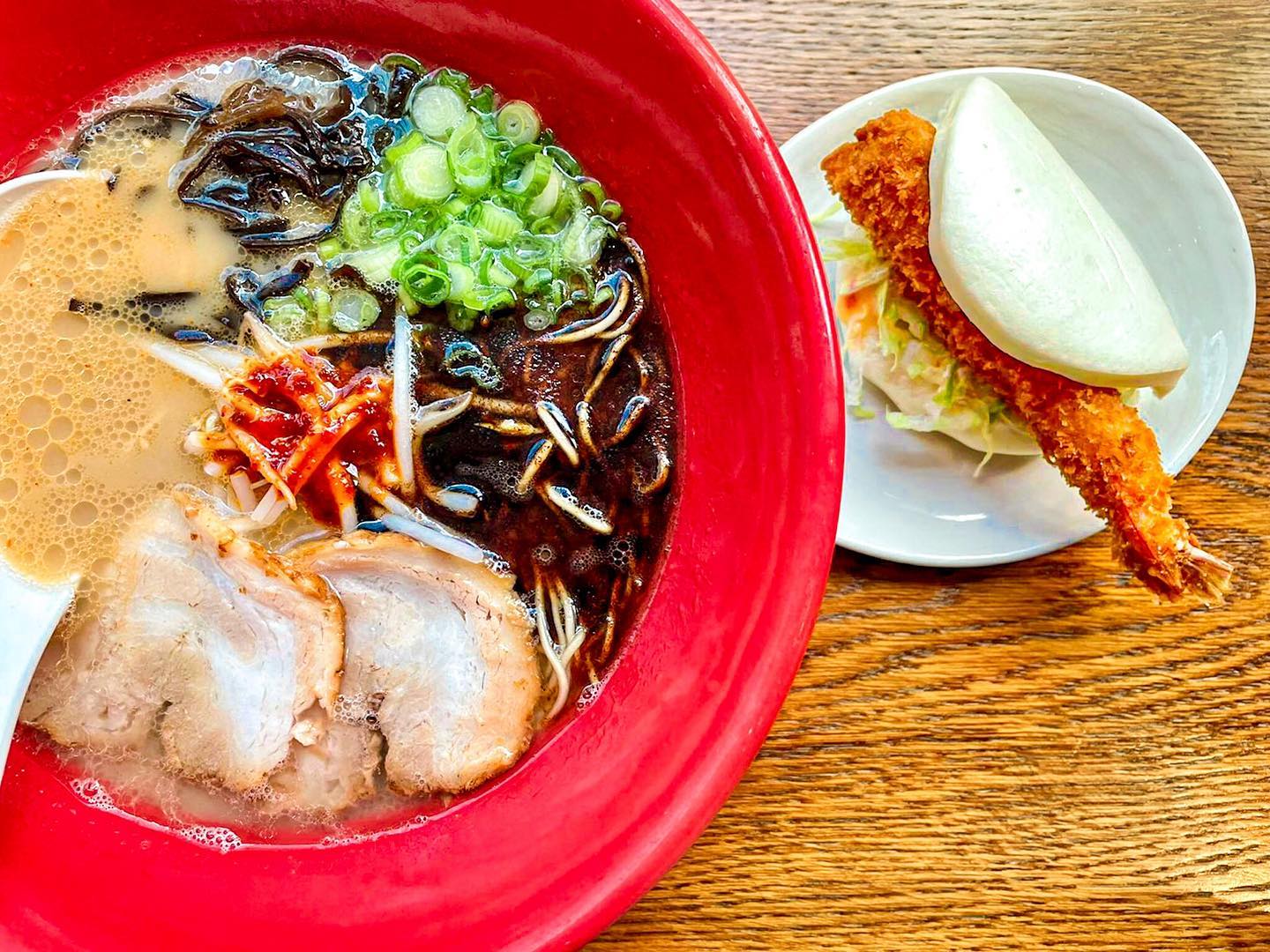 A bowl of ramen with sliced pork, green onions, bean sprouts, dark mushrooms, and a spicy sauce in a red bowl is placed next to a plate with a bao bun sandwich containing a crispy fried shrimp and some coleslaw, on a wooden table.