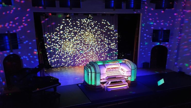 A stage with a brightly lit organ featuring colorful lights. The background features a curtain with a light projection resembling a starry night sky. There is also a grand piano on the left side of the stage. The venue is dimly lit with a focus on the organ.