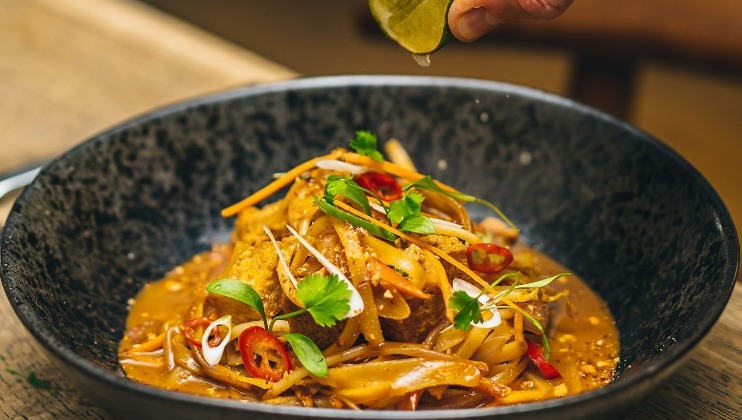 A hand is seen squeezing a lime wedge over a bowl of vibrant curry. The dish is garnished with fresh herbs, sliced red chilies, crispy vegetables, and a thick, flavorful sauce. The setting is a rustic wooden table, suggesting a cozy dining atmosphere.