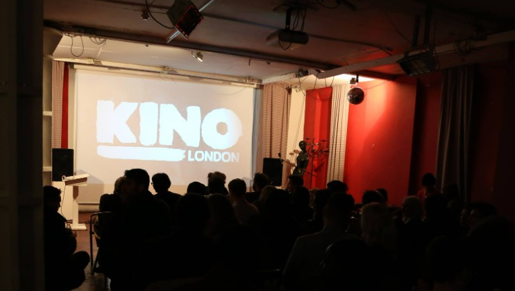 A dimly lit room filled with people seated and facing a large projection screen at the front. The screen displays the words KINO LONDON in bold white letters. The atmosphere suggests a film screening or presentation event.