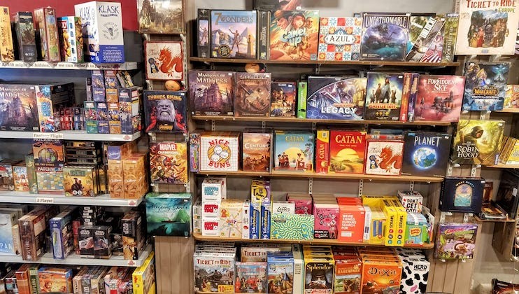 A vibrant display of various board games on shelves in a store. Games include Catan, Pandemic, Ticket to Ride, Azul, and others, with colorful boxes arranged neatly. The shelves are densely packed, showcasing a wide selection of games for different ages and interests.