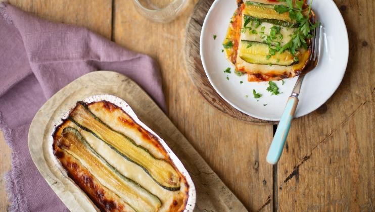 A serving of vegetable lasagna topped with fresh herbs on a white plate with a fork, next to a larger dish of lasagna placed on a wooden cutting board and a purple cloth, set on a wooden table.