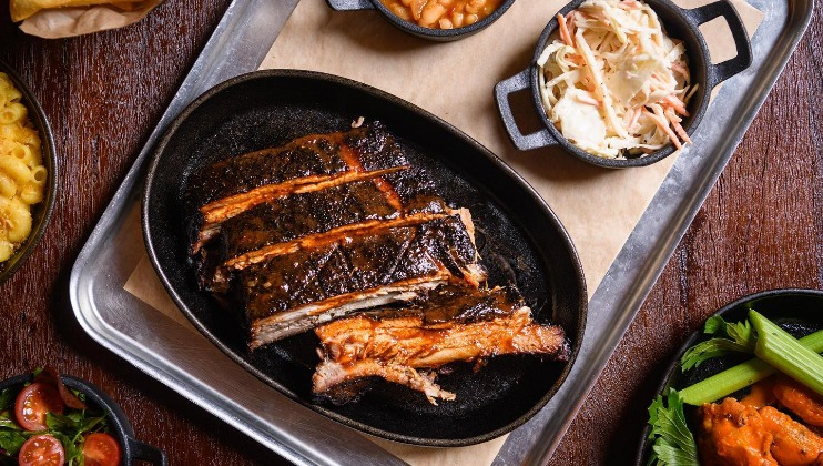 A platter of barbecued ribs served on a tray, surrounded by sides including a bowl of coleslaw, baked beans, celery sticks, macaroni and cheese, and a small salad with cherry tomatoes. The ribs are charred and arranged in a cast iron serving dish.