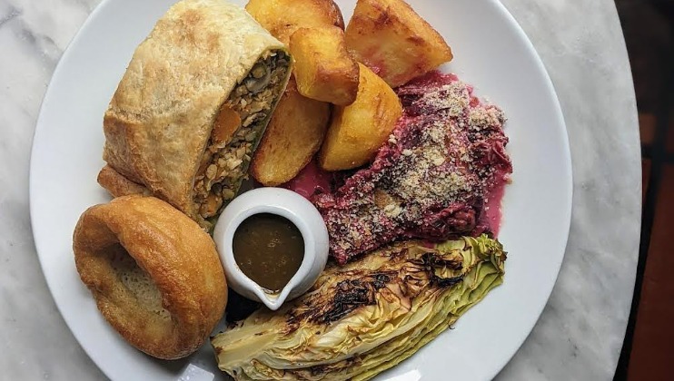 A white plate of food containing a serving of roasted vegetables wrapped in pastry, crispy roast potatoes, a helping of beetroot topped with cheese, caramelized roasted cabbage, a small jug of gravy, and a Yorkshire pudding.