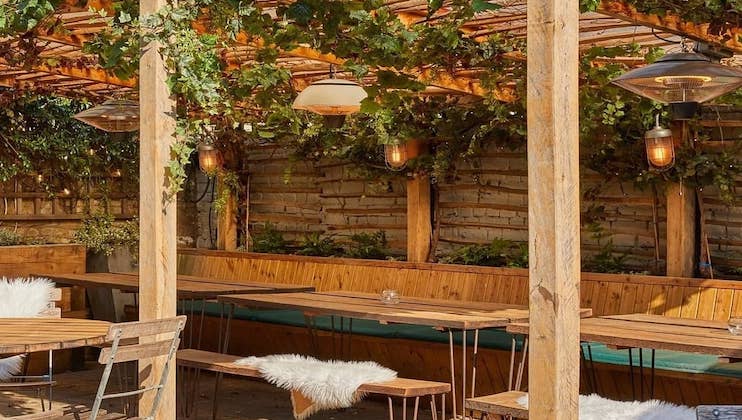 An outdoor seating area with wooden tables and benches beneath a pergola covered in green vines. String lights and hanging lamps provide warm illumination. Fur throws are draped over the benches for added comfort. Wooden fencing encloses the cozy setting.