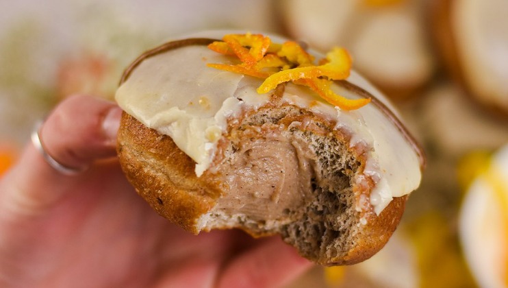 A close-up of a hand holding a doughnut with a bite taken out. The doughnut is filled with a creamy filling and topped with a white glaze and garnished with orange peel zest. The background is blurred, highlighting the doughnut as the main focus.