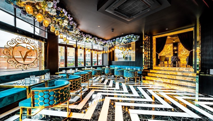 A luxury restaurant interior with a modern design, featuring blue velvet chairs, gold accents, and a black-and-white zigzag patterned floor. The ceiling is adorned with floral arrangements and decorative lighting fixtures. Large windows provide natural light.