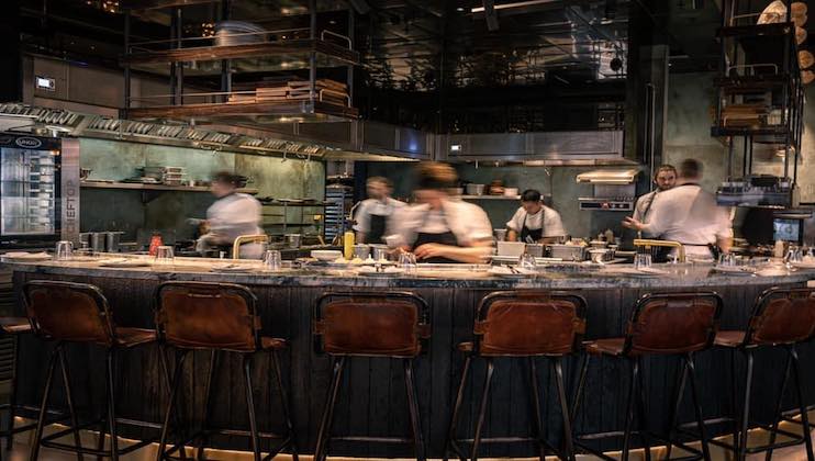 A modern, open-concept kitchen in a restaurant with chefs busy at work. The curved counter with a dark, rustic design is surrounded by several tall, leather-backed bar stools. The background displays various kitchen equipment and shelves laden with ingredients.