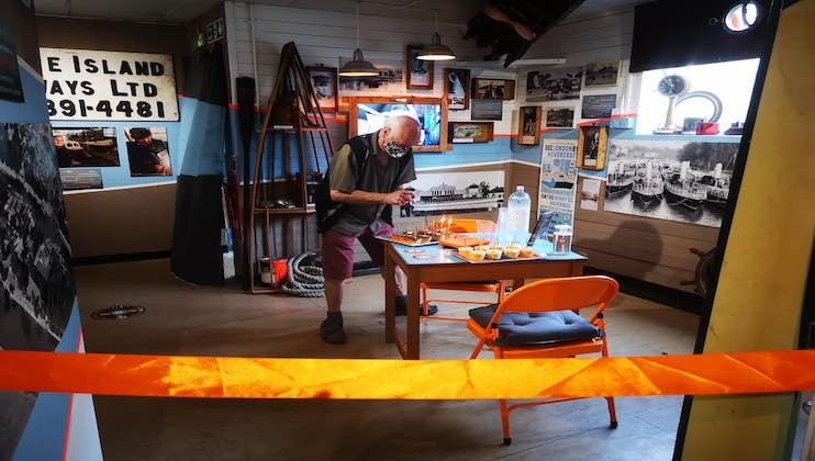 A person is standing inside a small, colorful museum or exhibition space, looking at items on a table. The space has numerous photographs and memorabilia on the walls. An orange chair and an orange barrier tape are also visible in the room.