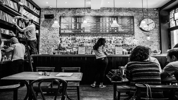 Black and white image of a cozy café with several people inside. One person is on a ladder, reaching for items on a high shelf, while others sit at tables or stand near the counter. A large menu board and a clock are visible on the brick wall behind the counter.