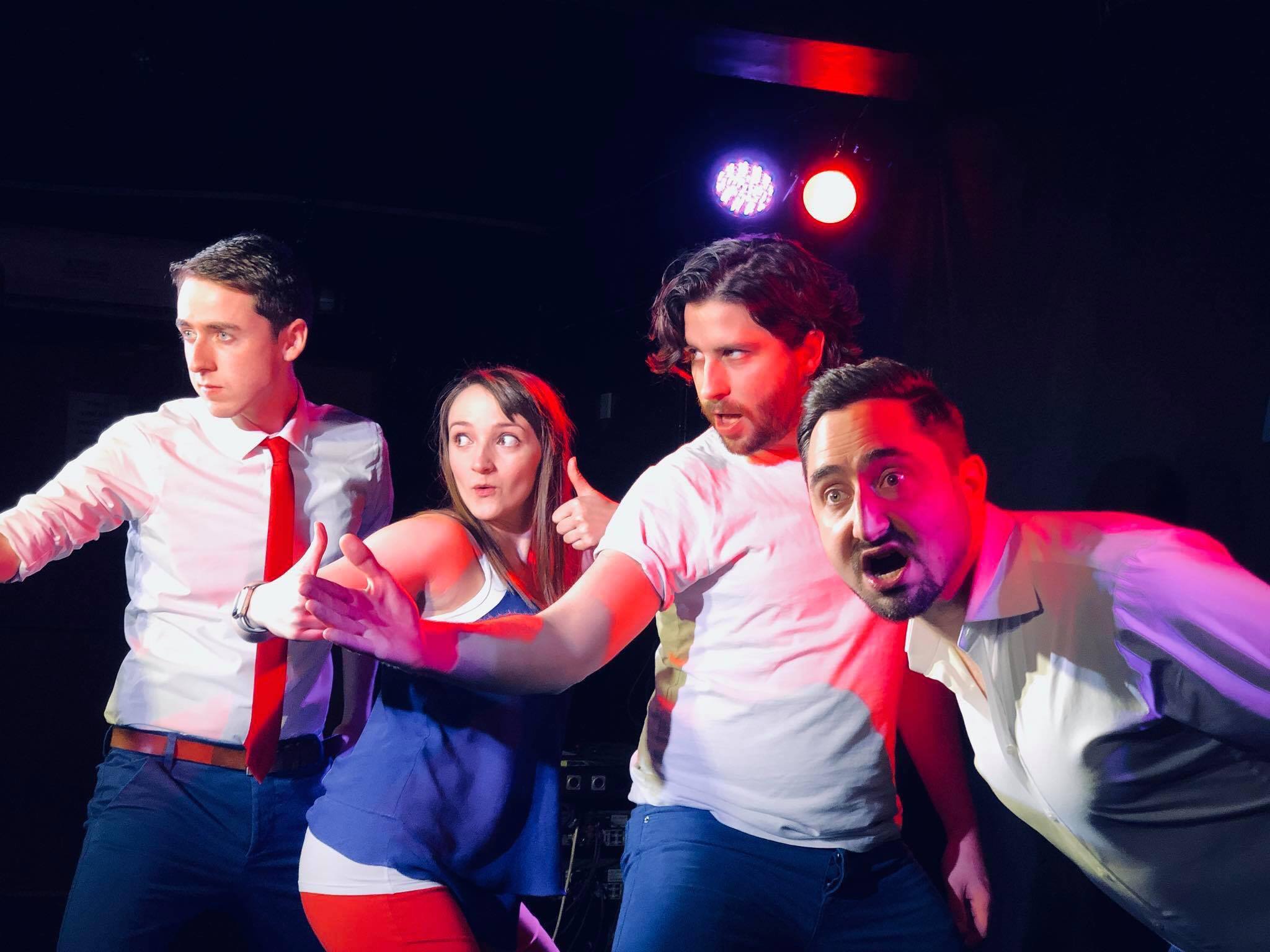 Four people on a dark stage with colorful lighting strike exaggerated, expressive poses. The man on the left points forward confidently, the woman next to him gestures dramatically, the third man raises his arm energetically, and the fourth looks surprised with his mouth open.