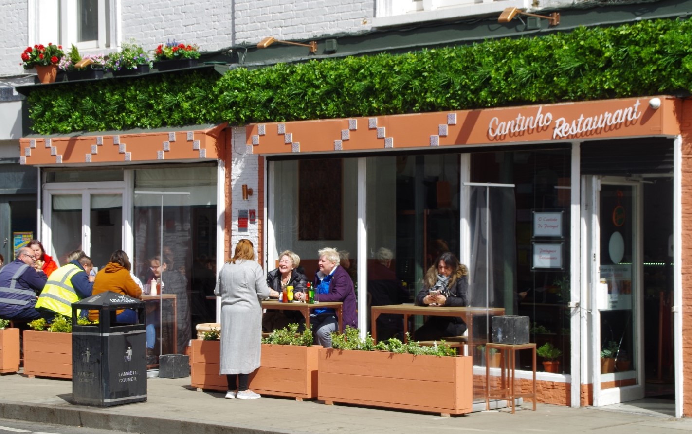 Street view of a restaurant called Canto do Restaurante. Several patrons are sitting at outdoor tables, eating and talking. The restaurant's exterior features an orange awning and signage, with green plants decorating the front. A person in a gray coat stands nearby.