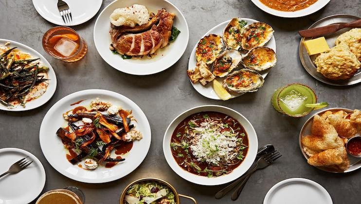 A table is filled with various dishes, including roasted meats, grilled vegetables, stuffed oysters, a bowl of rice with sauce, bread rolls, and vibrant drinks. The spread showcases a variety of flavors and textures, all beautifully presented on white and gold-rimmed plates.