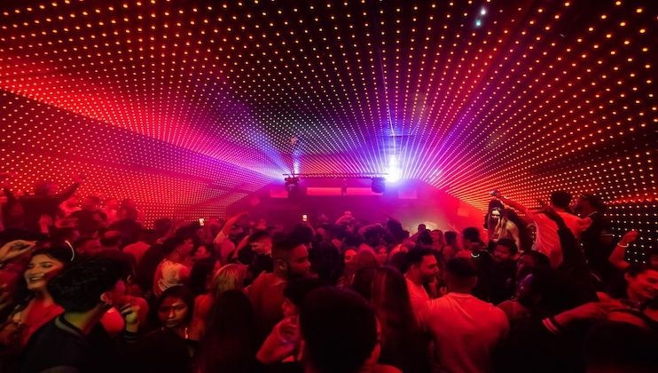 A crowded dance club with a vibrant light display on the ceiling. People are dancing and socializing amid red and purple lights radiating from a central point above the DJ booth. The atmosphere is energetic and lively.