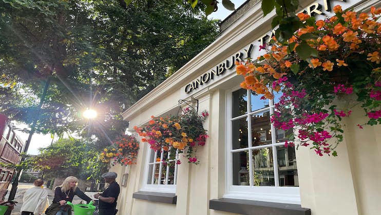 A sunlit street featuring a charming building with large windows adorned with vibrant flower baskets. Two people interact beside a green bin in front of the building. The sign above the door reads CANONBURY. Trees and additional buildings are visible.