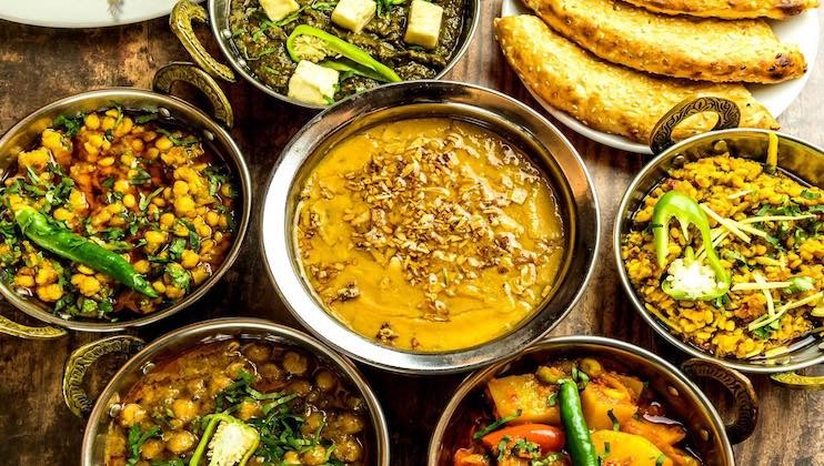 A variety of colorful Indian dishes served in metal bowls, includes lentils, vegetables, paneer, and curry, garnished with green chilies and herbs. Several pieces of naan bread are placed on a white plate in the background.