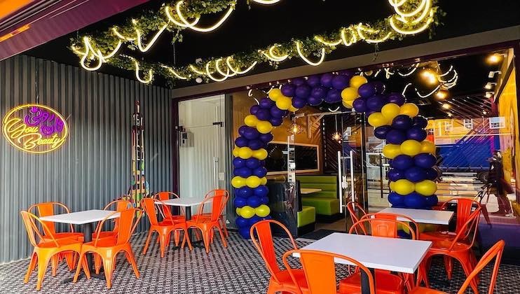 A brightly lit outdoor seating area features tables with orange chairs. A balloon arch in blue and yellow decorates the entrance to the adjacent building. String lights and greenery hang from the ceiling, and a neon sign on the left wall reads, Cool & Beauty.