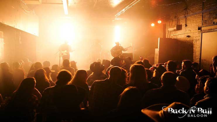 A crowd of people watches a live band perform on a dimly lit stage, illuminated by bright, warm lights. The silhouettes of the band members and audience are visible against the glow. In the bottom right corner, there is a logo that reads Buck'n Bull Saloon.