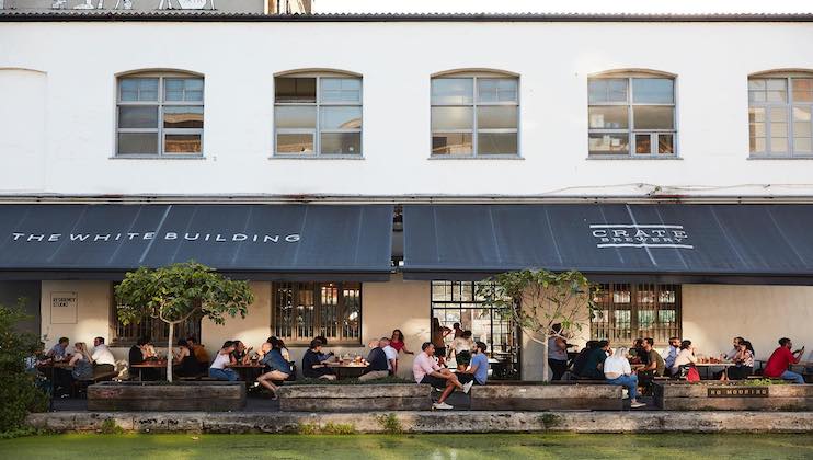 Outdoor view of people sitting and dining on long wooden benches under large black awnings with the text THE WHITE BUILDING and CRATE. The building has multiple large windows, and the setting appears to be a casual, social environment.