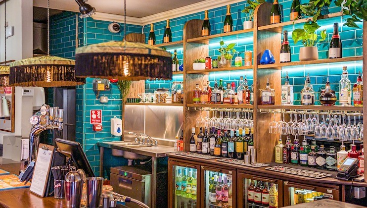 A vibrant bar with teal-tiled walls, wooden shelves filled with an assortment of liquor bottles and potted plants. The bar counter displays various bottles and glasses, while pendant lights with tassels hang from the ceiling, illuminating the cozy space.