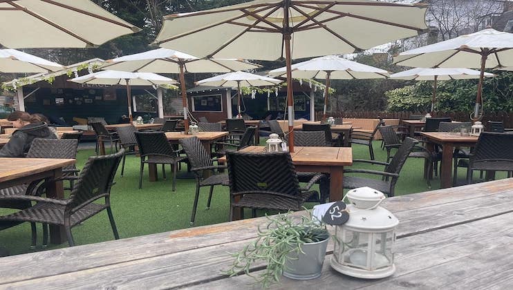 Outdoor dining area with wooden tables and wicker chairs, arranged under large beige umbrellas. A few decorative items, including white lanterns and small potted plants, are placed on tables. The area is surrounded by greenery and a rustic wooden fence.