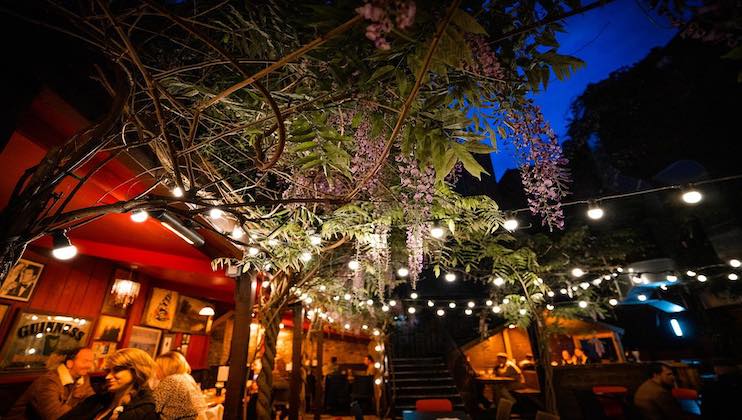 Outdoor restaurant patio at night, illuminated by string lights draped from trees with hanging purple flowers. Diners are seated at tables, enjoying their meals and conversations. The ambiance is lively and warm, complemented by a clear, dark blue sky.