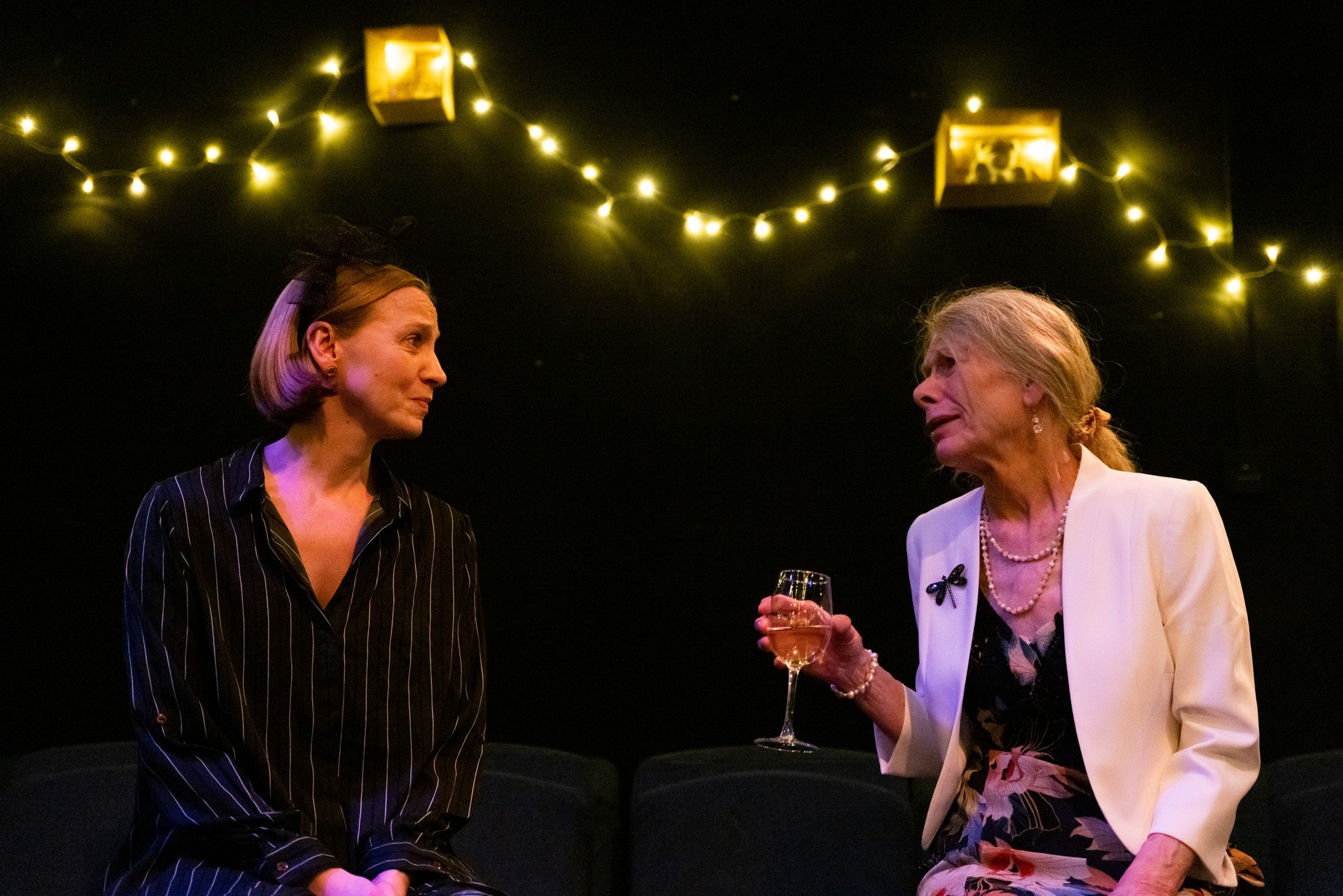 Two women are sitting and conversing. The woman on the left wears a dark, striped top with a headband. The older woman on the right is dressed in a white blazer over a floral dress, holding a wine glass. They appear to be on a stage set with string lights in the background.