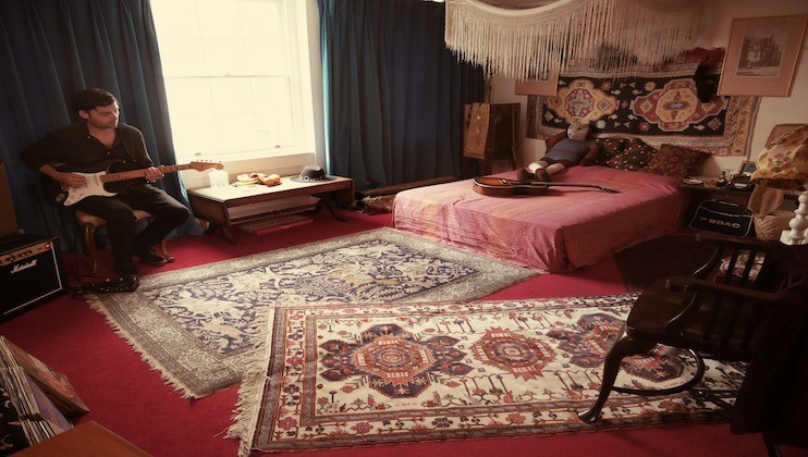 A person sits in a cozy room playing an electric guitar. The room is decorated with various rugs, tapestries, and a fringed canopy above the bed. A large window with blue curtains lets in natural light, illuminating the eclectic and artsy space.