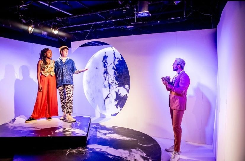 Three people in colorful outfits stand in a modern, abstract setting with white walls and black-and-white marble patterns. Two are on an elevated platform to the left, while one is holding a book to the right. Soft lighting illuminates the scene.