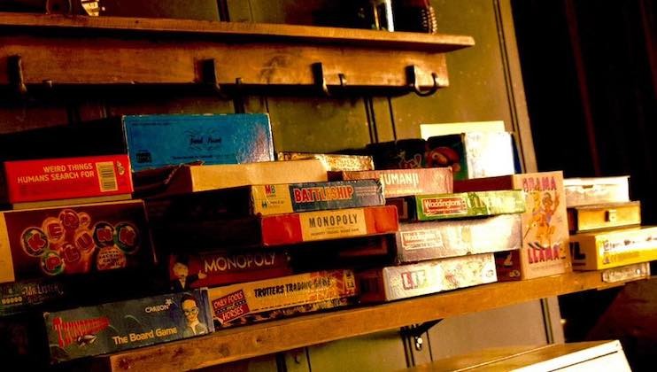 A shelf displaying a variety of board games, including Monopoly, Trivial Pursuit, Operation, and Battleship. The games are stacked in an organized yet casual manner, with the shelf situated against a dimly lit background.